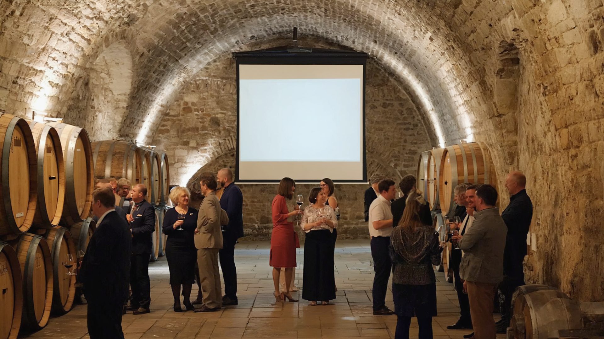 Guests standing together with glasses in a vaulted cellar; wooden casks and a small screen in the background.